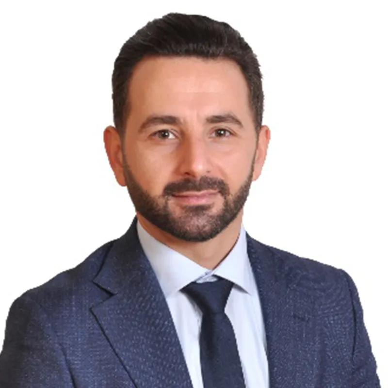 A professional headshot of a man with dark hair and a short, neat beard, wearing a blue patterned suit jacket, a light blue shirt, and a dark tie, set against a plain white background.