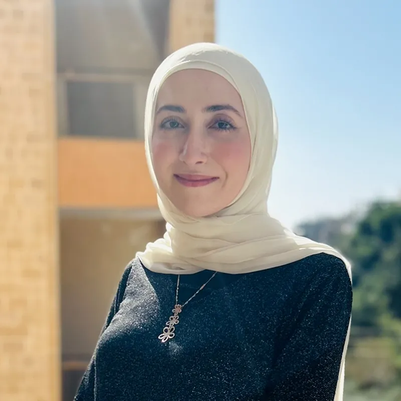 A portrait photo of a smiling woman wearing a light, creamy white hijab and a dark, sparkly top, standing outdoors on a sunny day with an out-of-focus beige building and blue sky in the background.