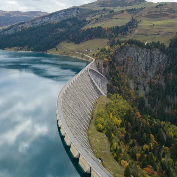 Aerial view of a massive, curved concrete arch-gravity dam holding a deep blue reservoir. The dam has a roadway running across its crest and is bordered on the right by a steep, rocky cliff and an evergreen forest with patches of autumn foliage.