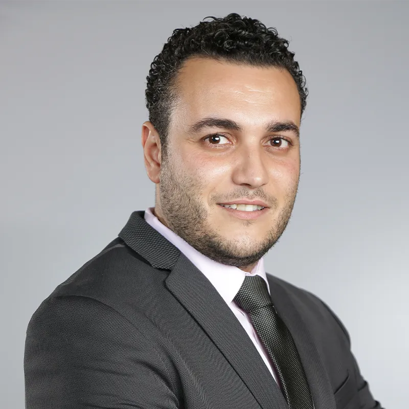 A headshot of a smiling man with dark, curly hair and a short beard, wearing a dark suit jacket, a light pink shirt, and a dark patterned tie, set against a plain gray background.