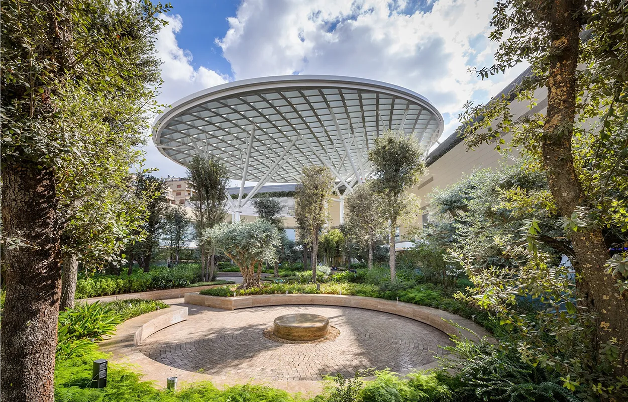 Street-level view of ABC Verdun Mall in Beirut showing the modern multi-story façade with glass panels, open terraces, and people walking along the ground-level shops and shaded walkways.