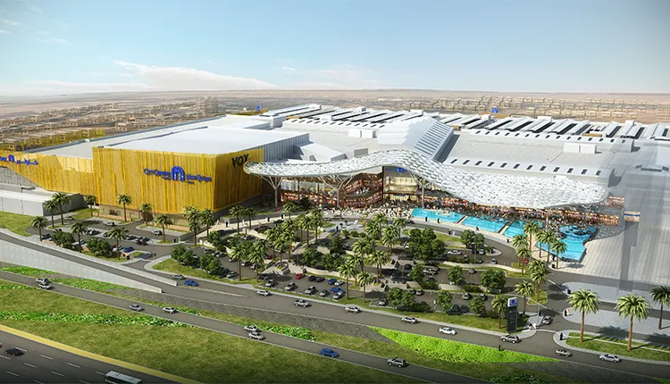 Architectural rendering of a large modern shopping mall with landscaped greenery, palm trees, outdoor seating areas, a swimming pool, and adjacent roadways with traffic; signage for VOX Cinemas and City Centre is visible on the building.