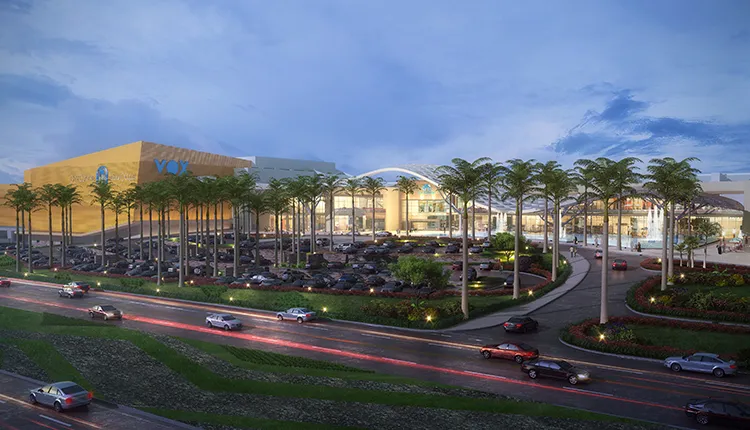Evening architectural rendering of a large shopping mall with illuminated VOX Cinemas signage, a spacious parking lot surrounded by palm trees, landscaped gardens, and cars on adjacent roads under a partly cloudy sky.