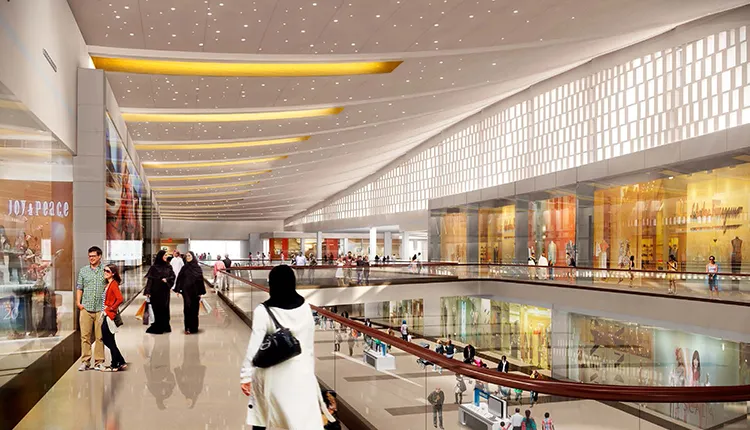 Interior view of a modern shopping mall with high ceilings, large windows, multiple floors, and people walking and shopping, including women in traditional Middle Eastern attire.