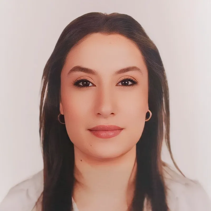 A professional passport-style headshot of a young woman with long, dark hair, wearing a white top and small hoop earrings, looking directly at the camera against a plain light background.