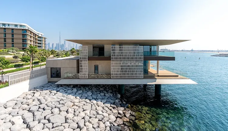 Modern waterfront villa with large overhanging roof and patterned screens, built on stilts above rocky shore, overlooking calm blue water with a city skyline in the background.