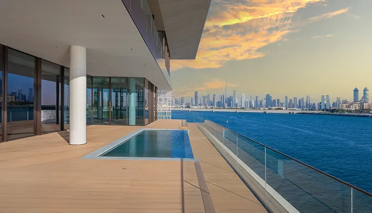 Modern balcony with a small pool overlooking a large body of water and a distant city skyline under a colorful sunset sky.