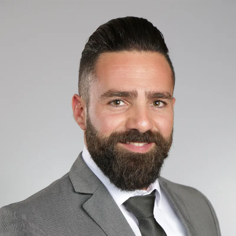 A headshot of a smiling man with dark hair styled back, a full, dark beard, wearing a light gray suit jacket, white shirt, and a dark patterned tie, set against a plain gray background.
