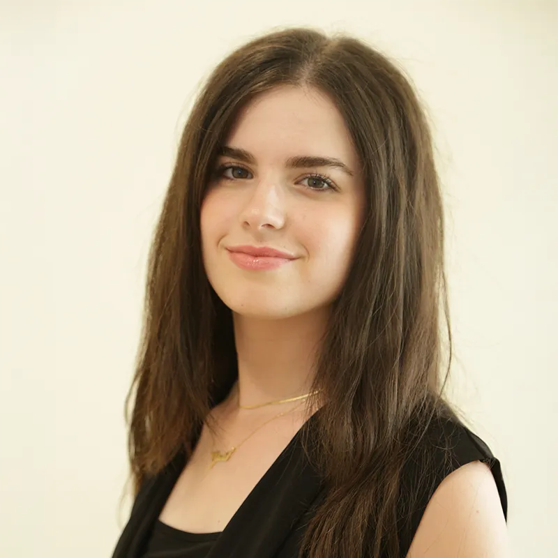 A headshot of a young woman with long, dark hair, wearing a black top and a delicate gold necklace, smiling subtly and looking toward the camera, set against a plain, light background.