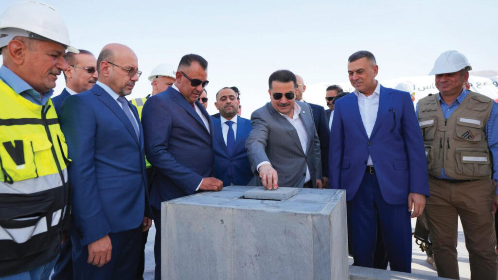 Group of officials and workers at a construction site, a man in a gray suit places a concrete block as others watch the ceremony.